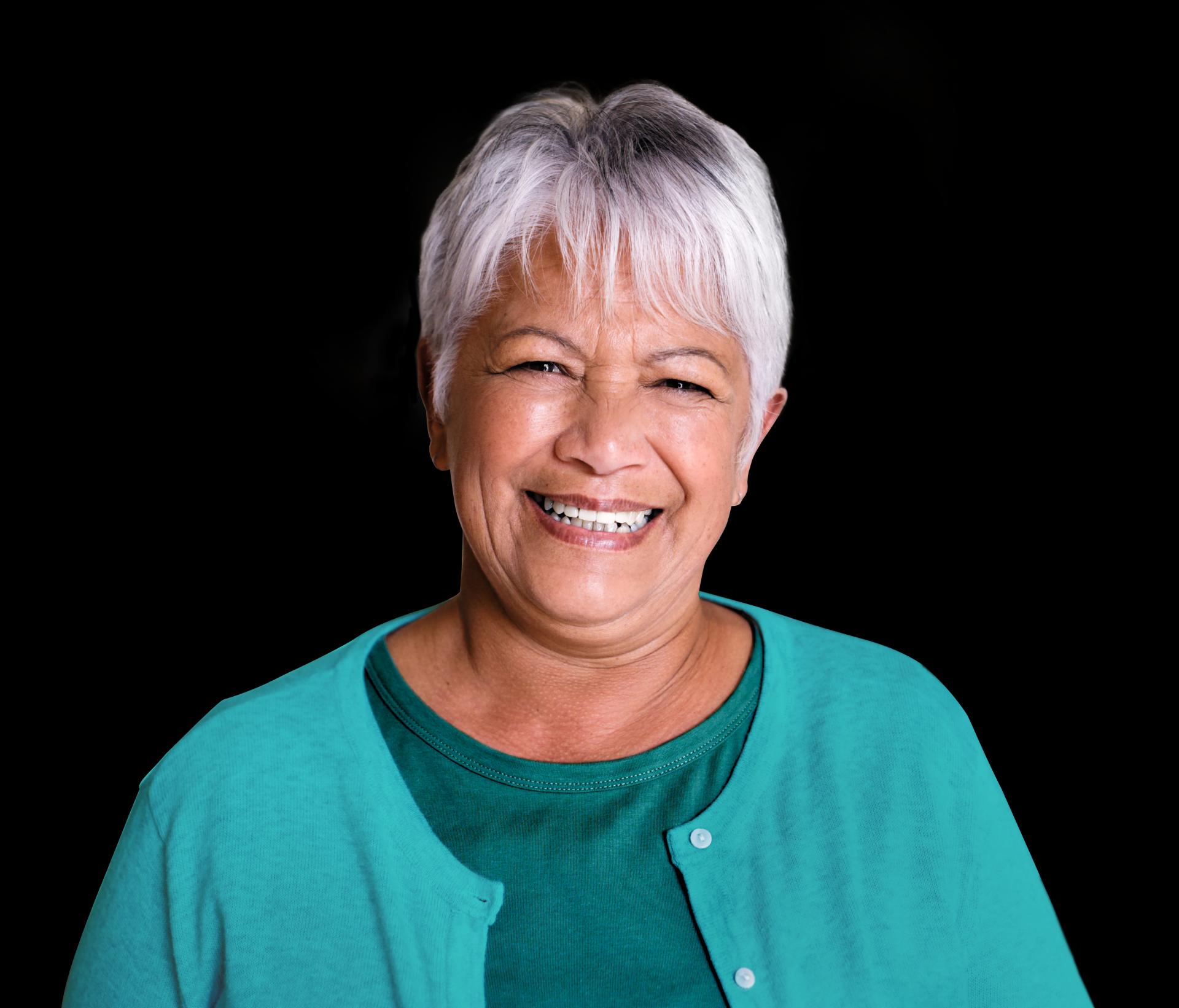 Mulher jovem de cabelos curtos e loiros sorrindo amplamente, vestindo um moletom verde e um relógio azul no pulso. Ela aponta para suas bochechas com ambos os dedos indicadores, transmitindo alegria e positividade.